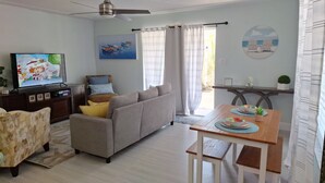A living room with a sofa, a television, a dining table with placemats, and a ceiling fan. - BEACHSIDE COZY GETAWAY! Walk/Bike to Beach,Dining,Bars,Small fenced yard for pet (Naples)