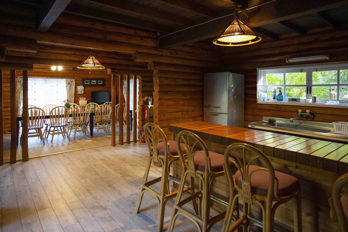 A rustic wooden cabin interior with a dining area, wooden chairs, a bar with stools, a refrigerator, and a window with a view of greenery.