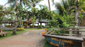 Entrada a un resort tropical con un letrero que dice 'Welcome to Blue Wave', una hamaca y exuberante follaje.
