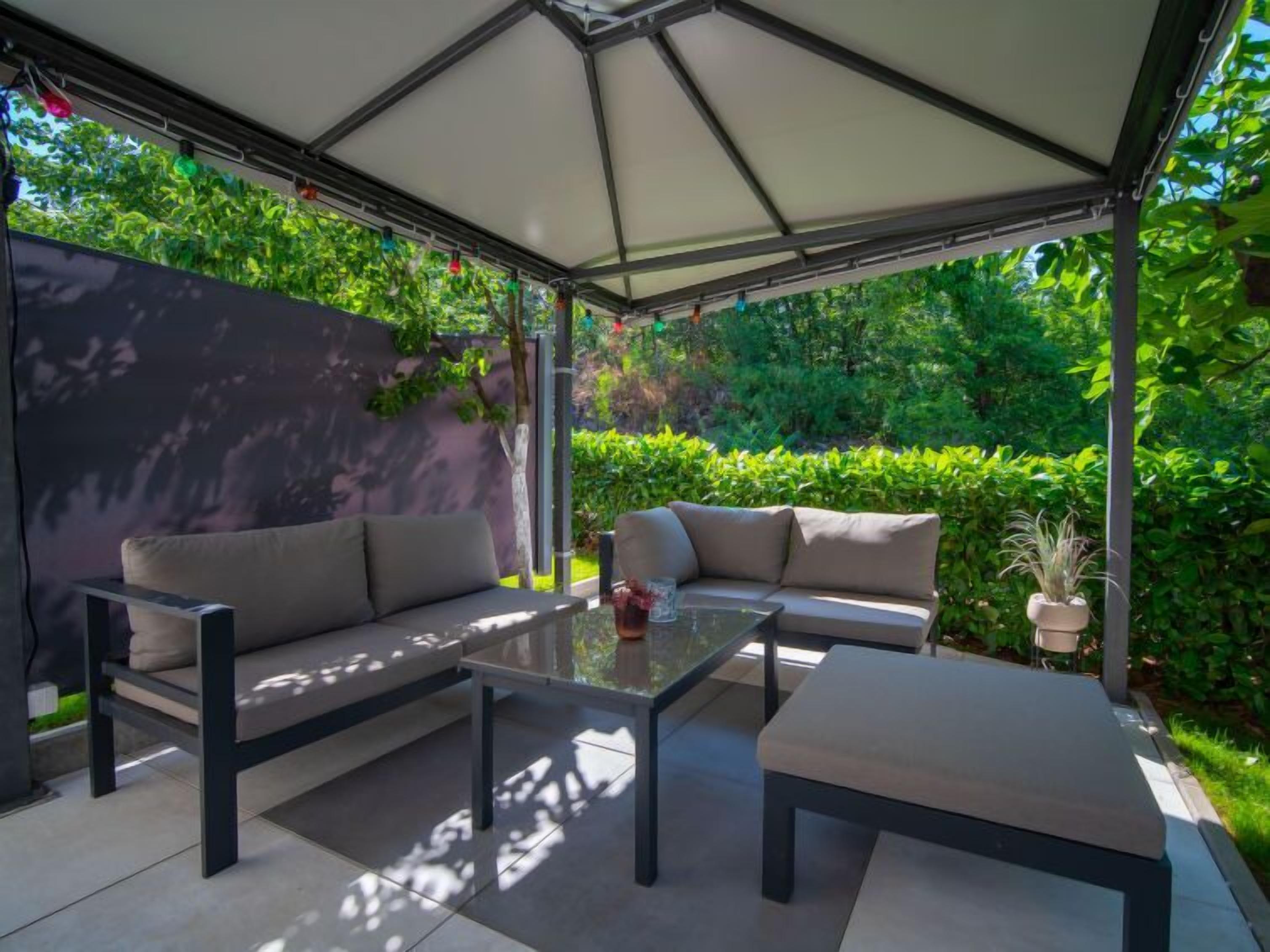 An outdoor patio area with a covered seating arrangement, including a sofa and a table, surrounded by lush greenery.