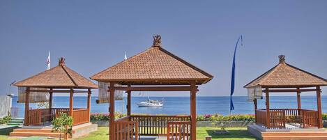 Tres gazebos de madera con techos de teja, cada uno con un banco y una pequeña mesa, ubicados en una zona cubierta de césped con vistas al mar y un barco al fondo.
