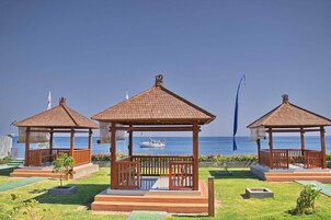Três gazebos de madeira com telhados de azulejo, cada um com um banco e uma pequena mesa, dispostos numa área gramada com vista para o mar e um barco ao fundo.