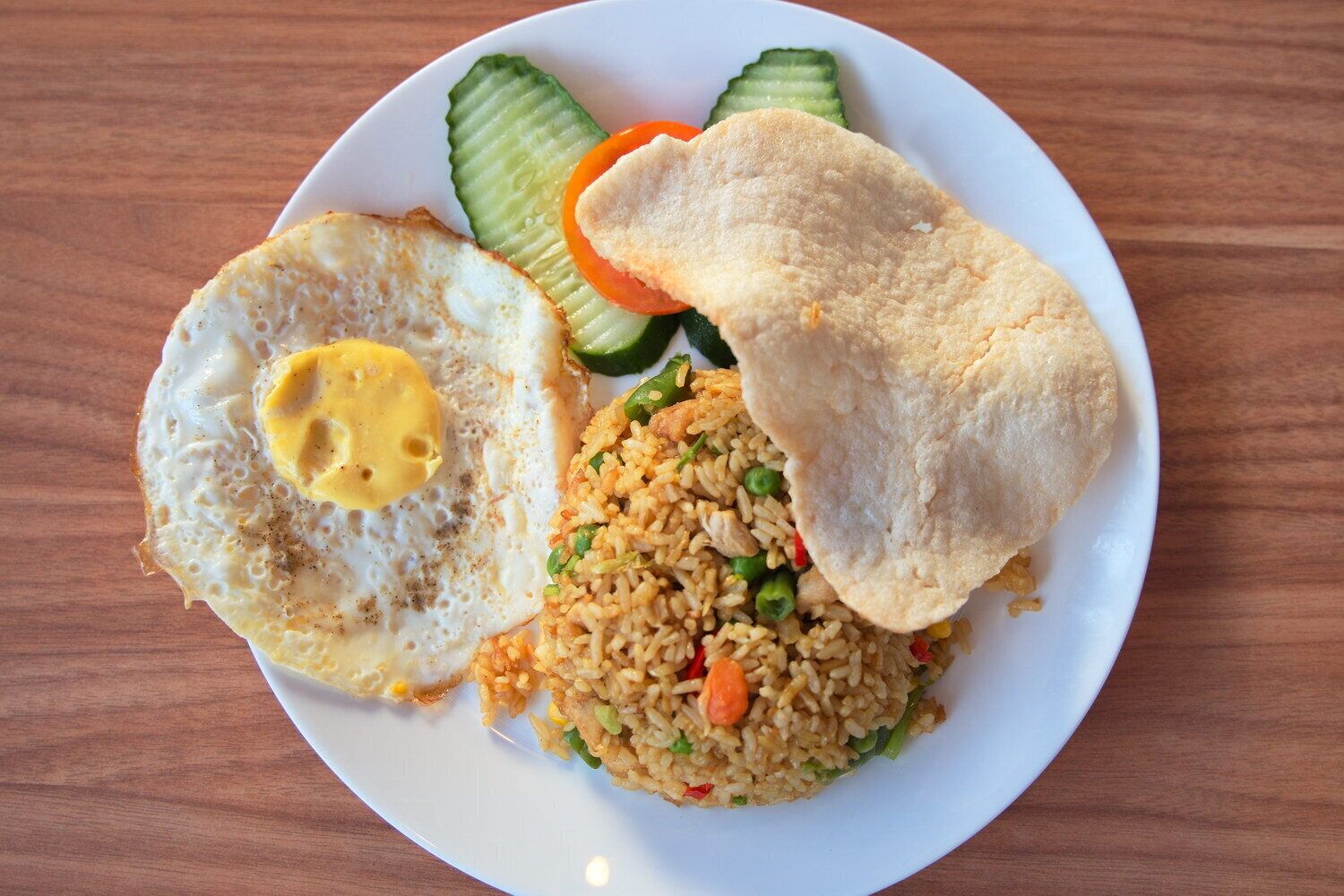 A plate with fried rice, a fried egg, cucumber slices, carrot slices, and a piece of bread.