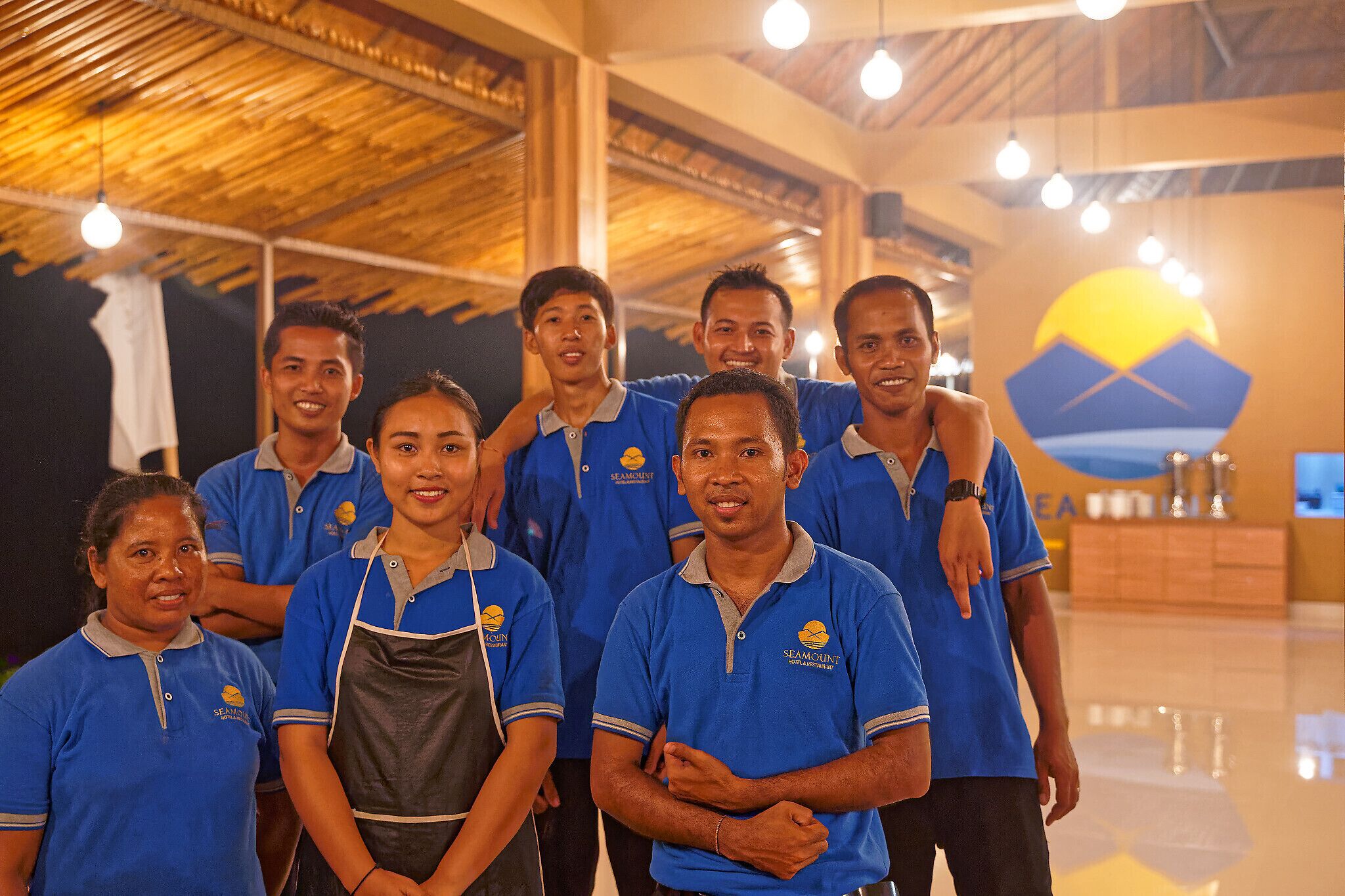 A group of people in blue shirts with a logo, standing in a spacious indoor area with a high ceiling and a large decorative wall.