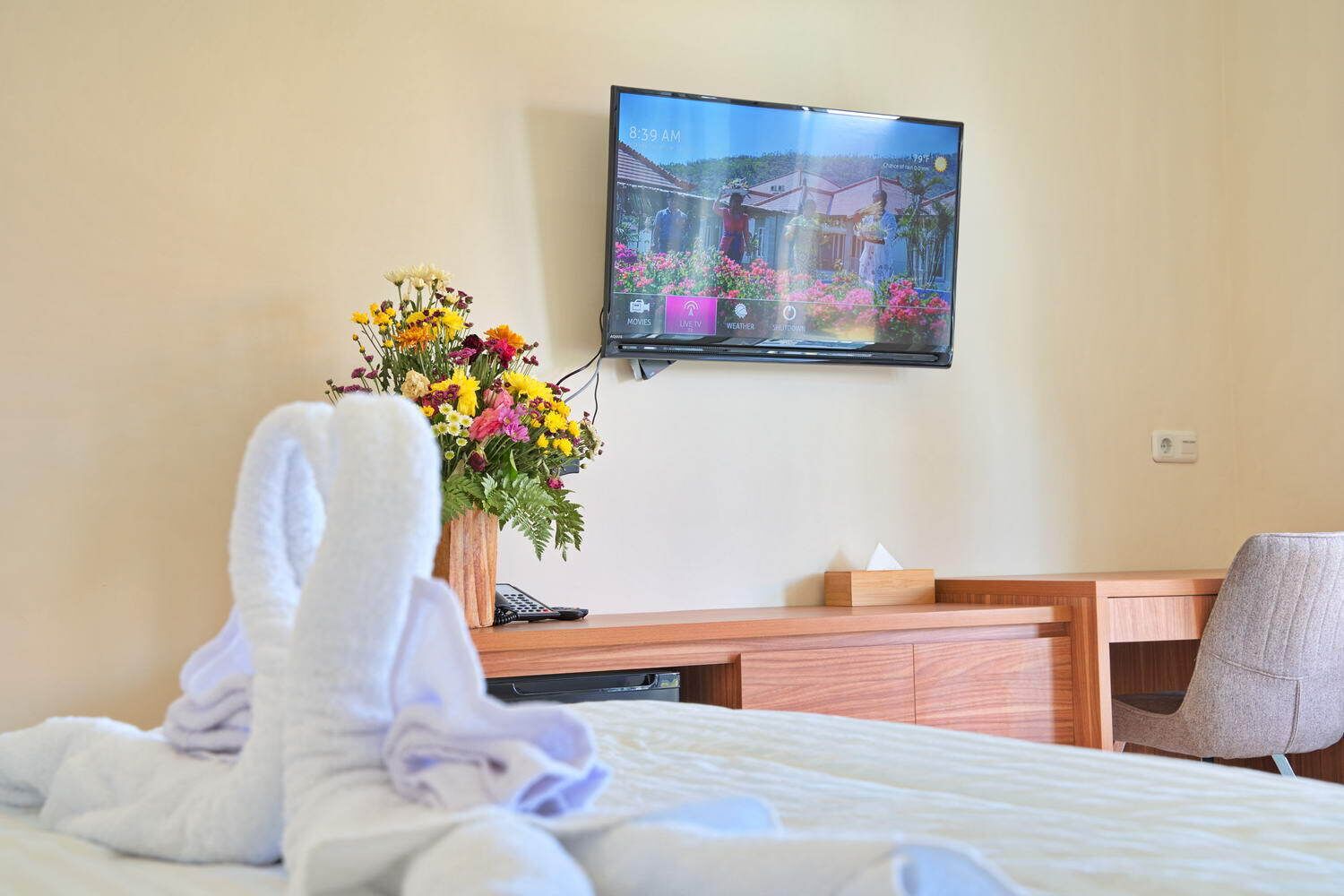 A hotel room with a bed, a desk, a chair, a TV mounted on the wall, and a vase of flowers on the desk.