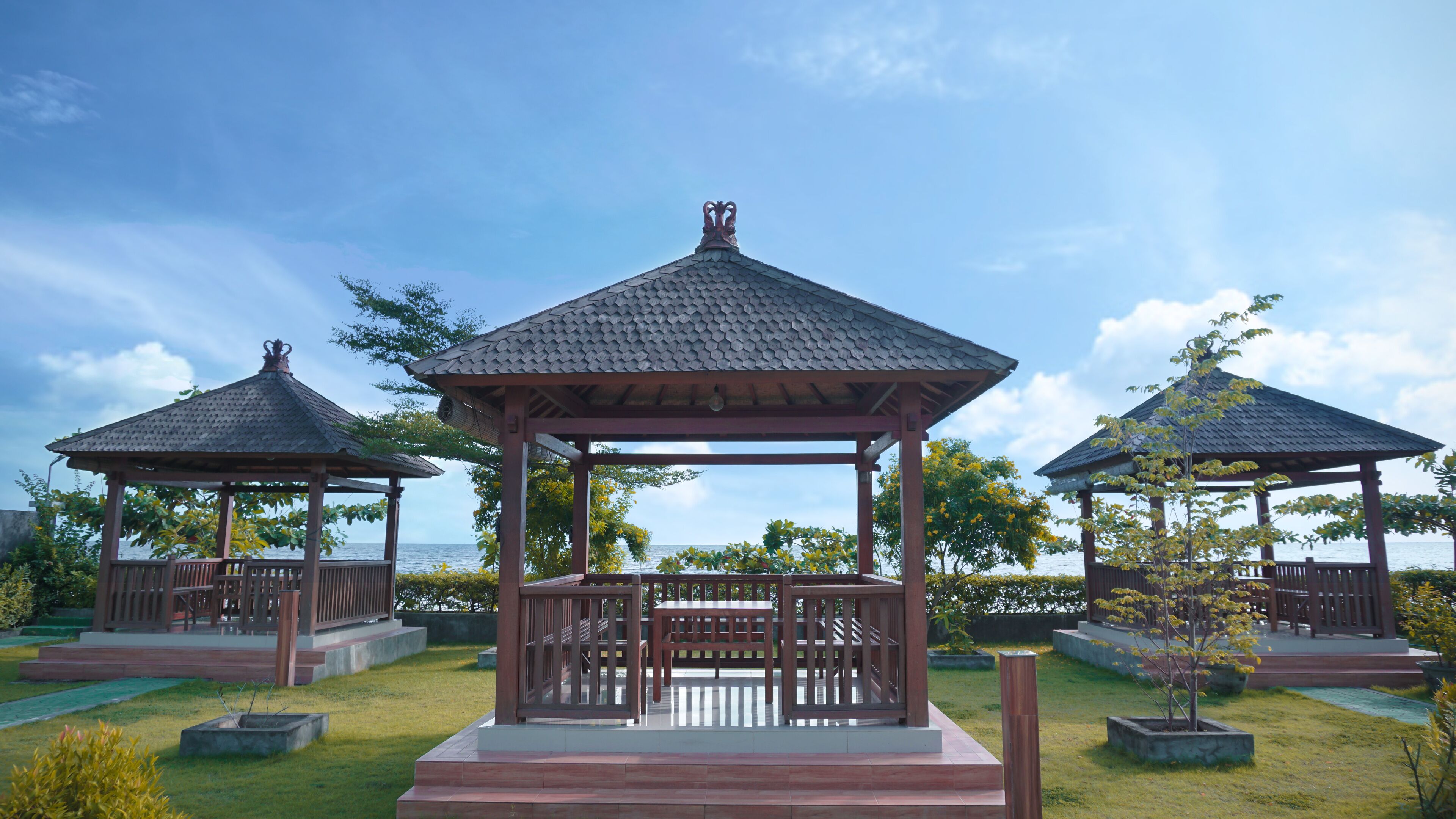 A gazebo with a tiled roof and a bench in the center, surrounded by a grassy area and trees.