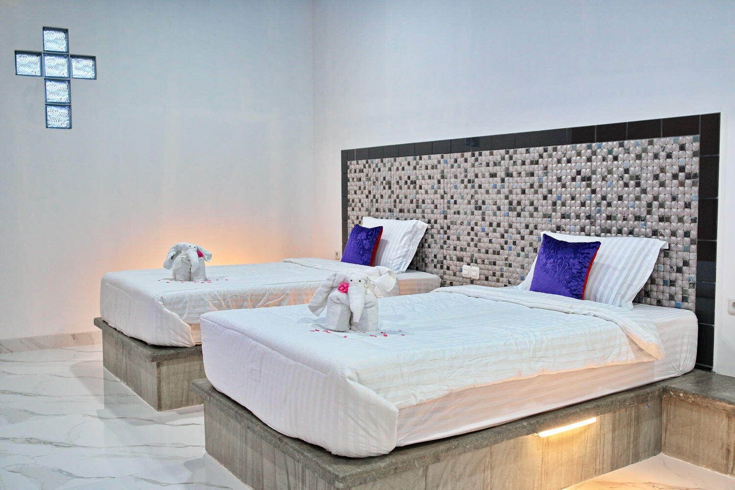 Two beds with white linens, a textured headboard, and a cross on the wall.