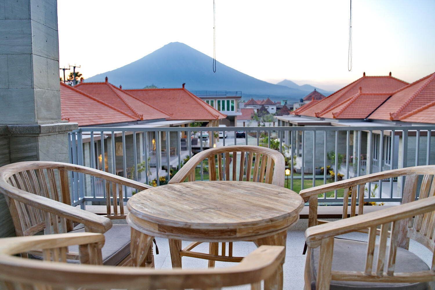 A balcony with wooden chairs and a table, overlooking a town with red-roofed buildings and a mountain in the background.