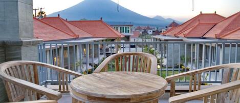 A balcony with wooden chairs and a table, overlooking a town with red-roofed buildings and a mountain in the background.