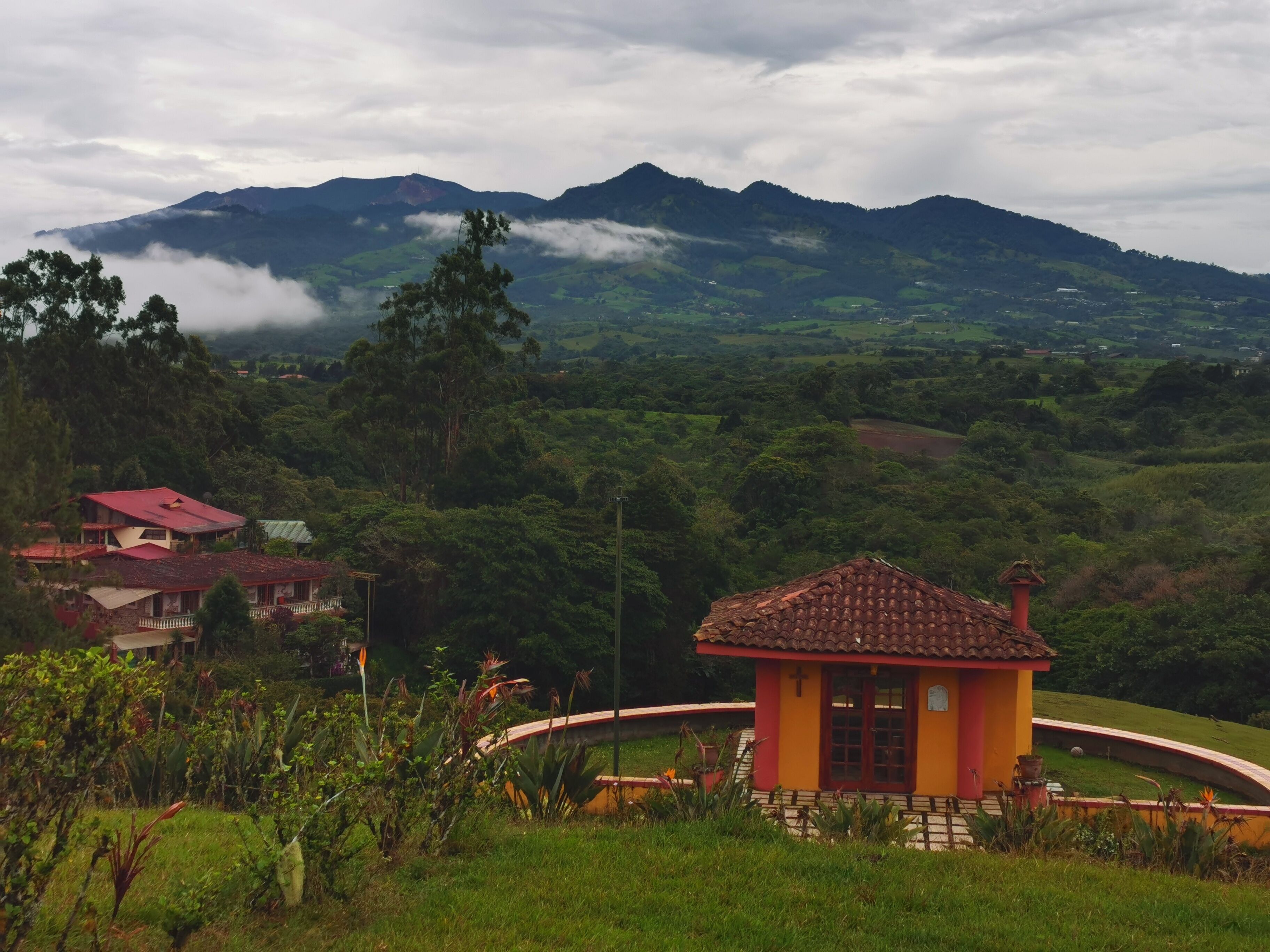 A small, yellow building with a red roof, surrounded by lush greenery and a mountainous backdrop.