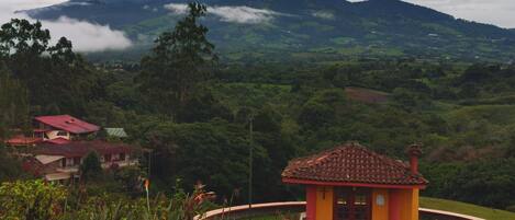 A small, yellow building with a red roof, surrounded by lush greenery and a mountainous backdrop.