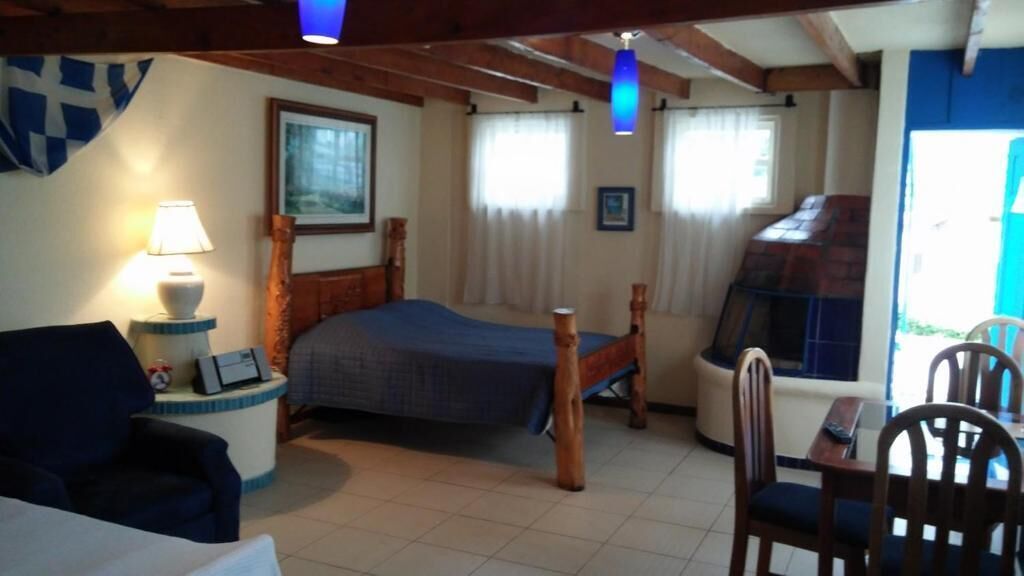 A bedroom with a bed, a bedside table with a lamp, a chair, a wooden headboard, and a window with curtains.
