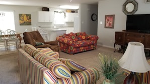 A living room with a sofa, a patterned armchair, a floral couch, a dining area with chairs, a kitchen area with cabinets, a clock, and a television. - Lazy Daze! Pups OK, Ideal location! Fenced Yard, Nice House. Wonderful! (Gulf Shores)