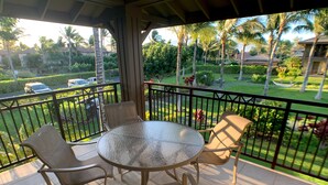A covered outdoor area with a round table and four chairs, overlooking a well-maintained garden with palm trees and a view of a residential area.