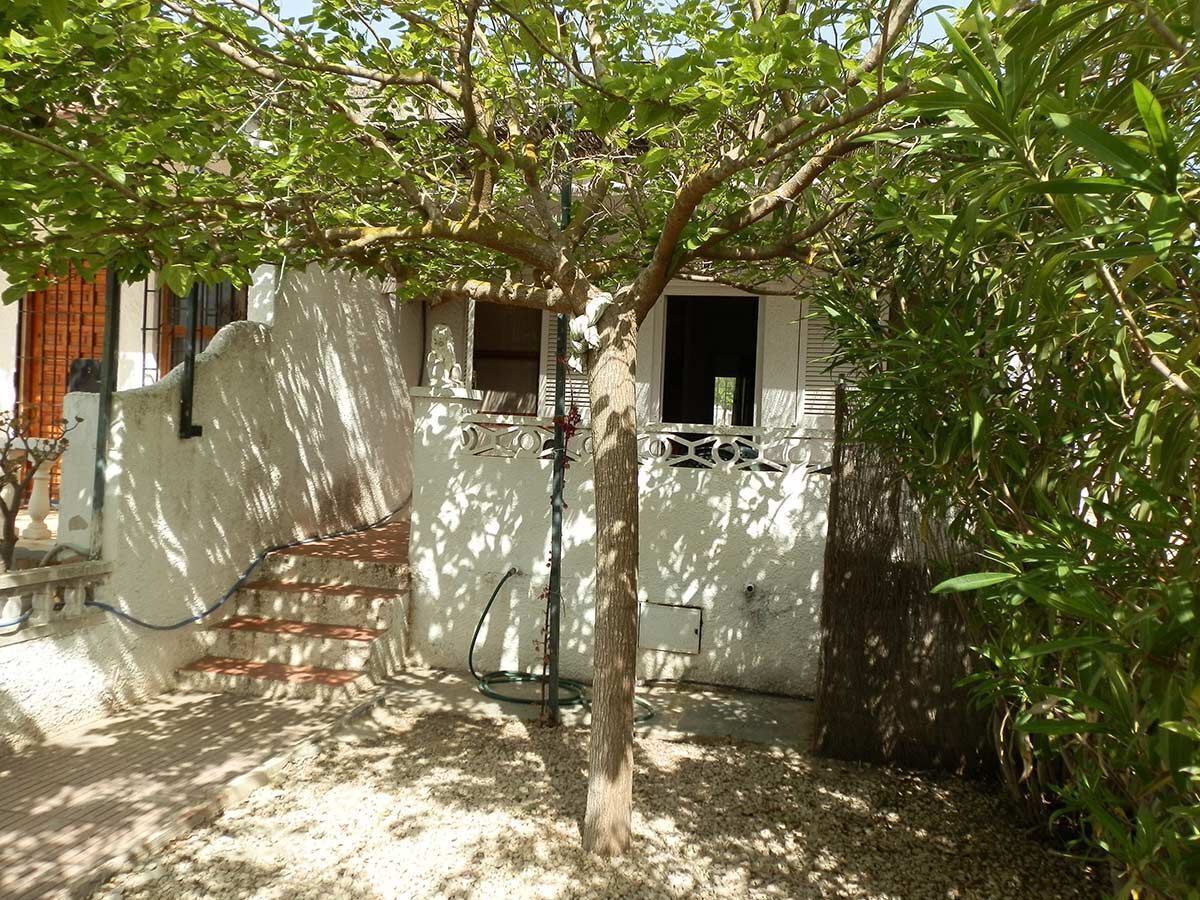 A white building with a staircase leading to a door, surrounded by greenery and a tree.