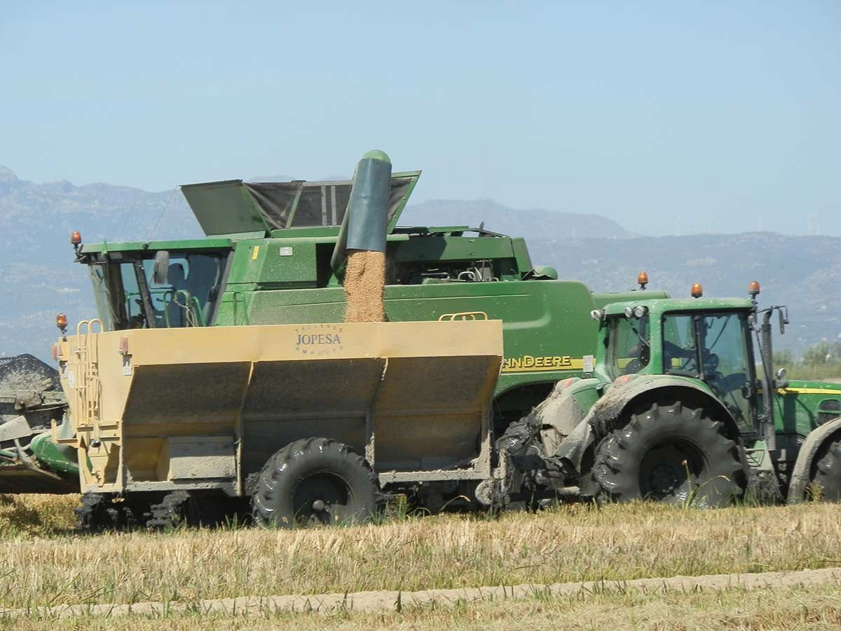 A green combine harvester with a yellow front loader attached, parked in a field with mountains in the background.