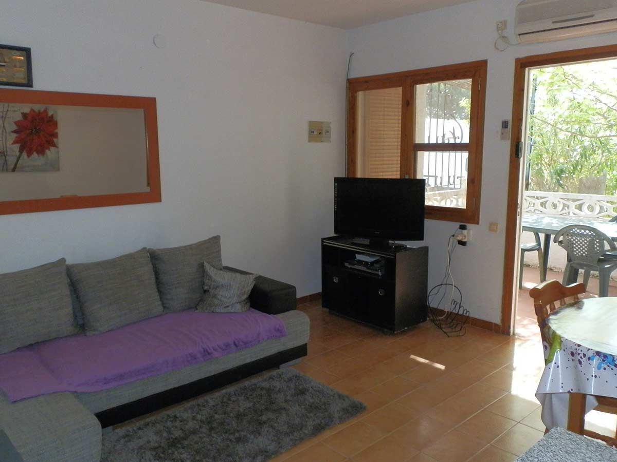 Een woonkamer met een bank, een tv op een dressoir, een eettafel met stoelen en een raam met zonwering.