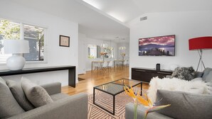 Ein modernes Wohnzimmer mit einem Glastisch, grauen Sofas, einem Essbereich mit Holztisch und Stühlen sowie einem großen Gemälde an der Wand.