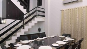 Un coin repas avec une longue table dressée pour six personnes, des chaises noires et un escalier moderne en arrière-plan.
