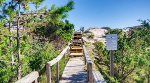 A wooden boardwalk leading to a beach, surrounded by pine trees and a sign indicating shared access with a house.