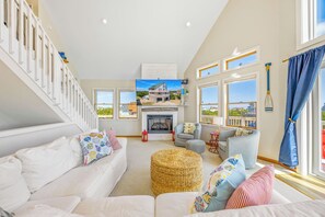 Een ruime woonkamer met een witte bank, een open haard en een groot raam met uitzicht op een strand.