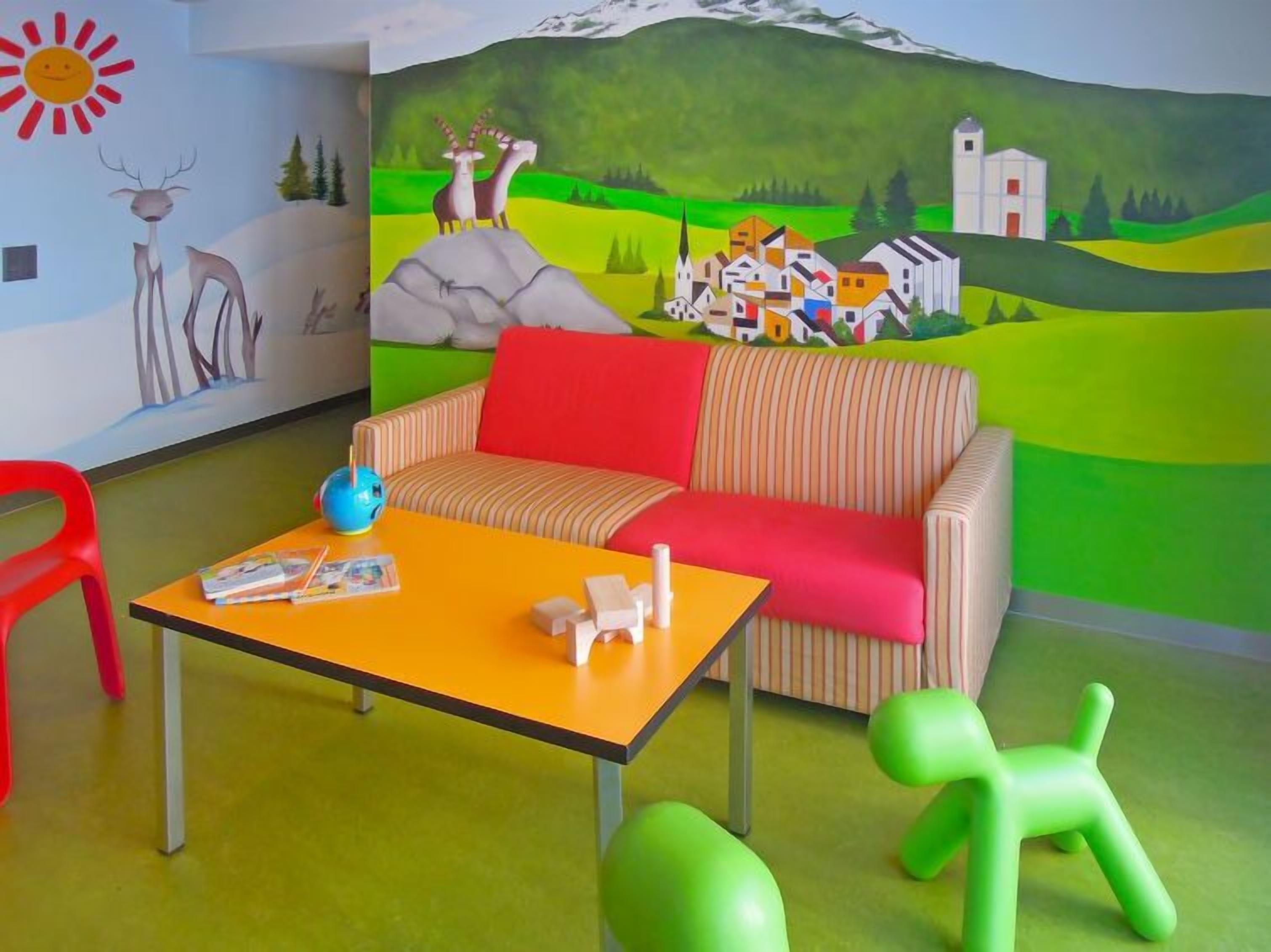 Une pièce avec un canapé à rayures, une petite table sur laquelle se trouvent un globe terrestre et des livres, un fauteuil rouge et une fresque murale représentant un paysage de village.
