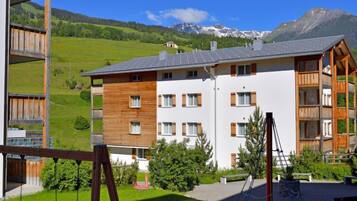 Un immeuble à plusieurs étages, dont les façades sont composées d’un mélange de bois et de murs blancs, se dressant sur un fond de collines et de montagnes verdoyantes.