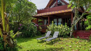 A two-story wooden house with a balcony, surrounded by lush greenery and a small lawn with two lounge chairs. - Thai Style Wooden Beachfront Villa with Private Sala (Koh Samui)