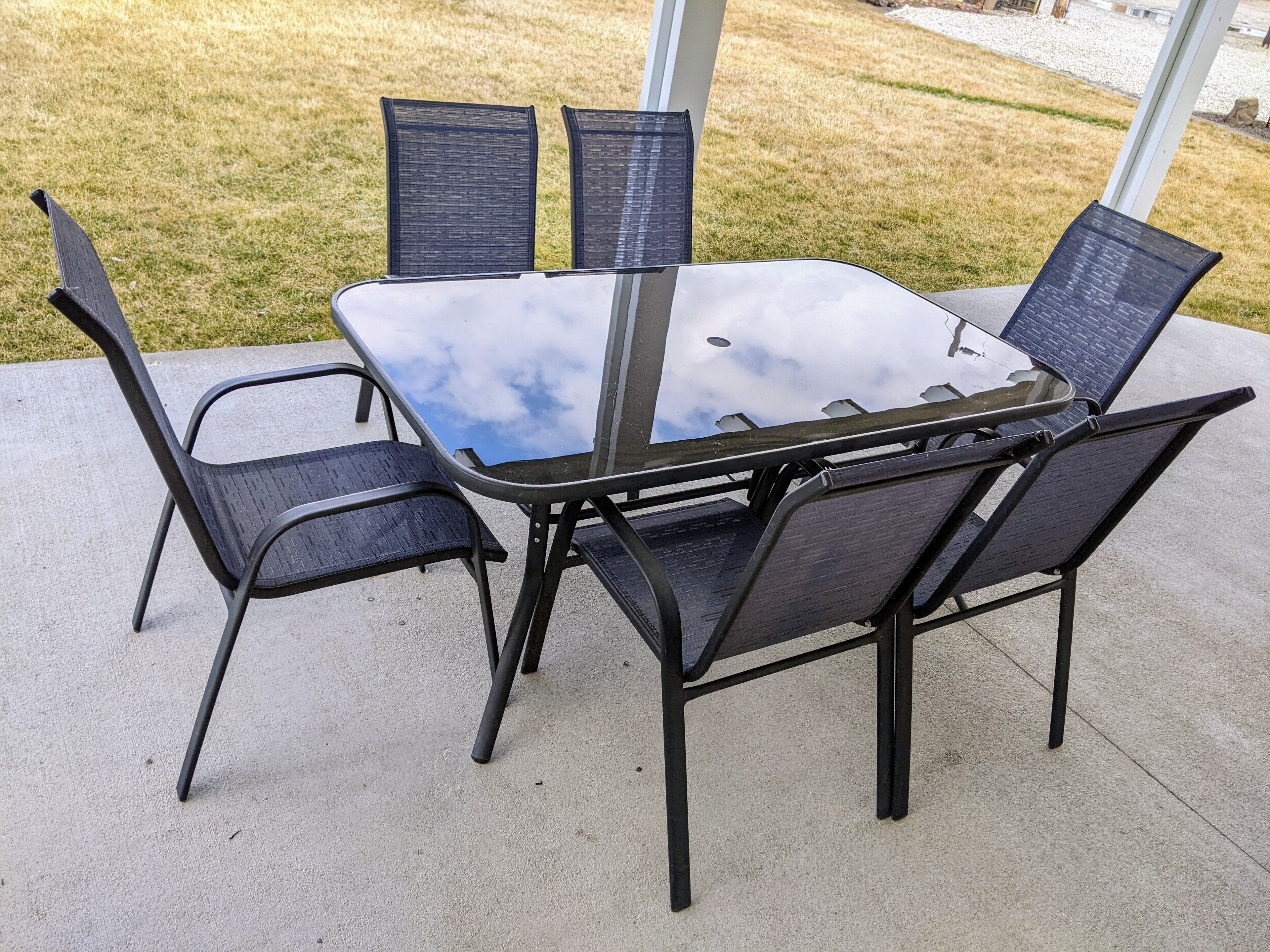 A glass-top table with four chairs, set on a concrete patio with a grassy area in the background.