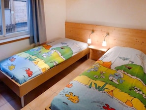 Ein Zimmer mit zwei Einzelbetten, jeweils mit farbenfroher Bettdecke im Cartoon-Design. Es gibt zwei Nachttische mit Lampen und ein Fenster mit Vorhängen.