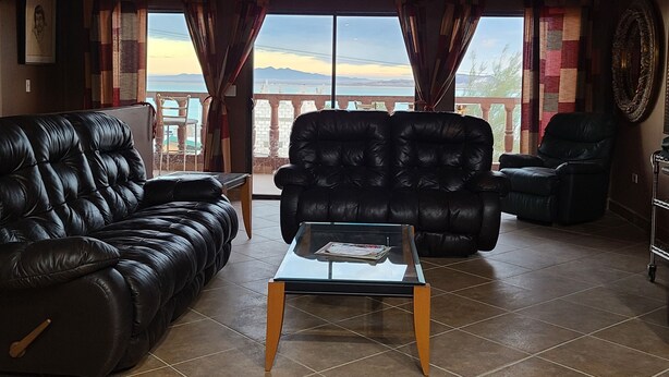 Een woonkamer met een zwarte leren bank, een zwarte lederen fauteuil en een glazen salontafel. Gordijnen en een uitzicht op een berglandschap door grote ramen.