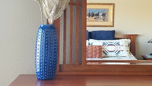 Eine Holzkommode mit einer Vase mit getrocknetem Pampasgras, einem gerahmten Bild eines Pferdes und einem Bett mit gemusterten Betttextilien.