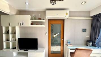 A compact living space with a flat-screen TV, a desk, a chair, a dining table, and a built-in shelf unit.