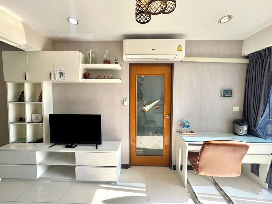 A compact living space with a flat-screen TV, a desk, a chair, a dining table, and a built-in shelf unit.