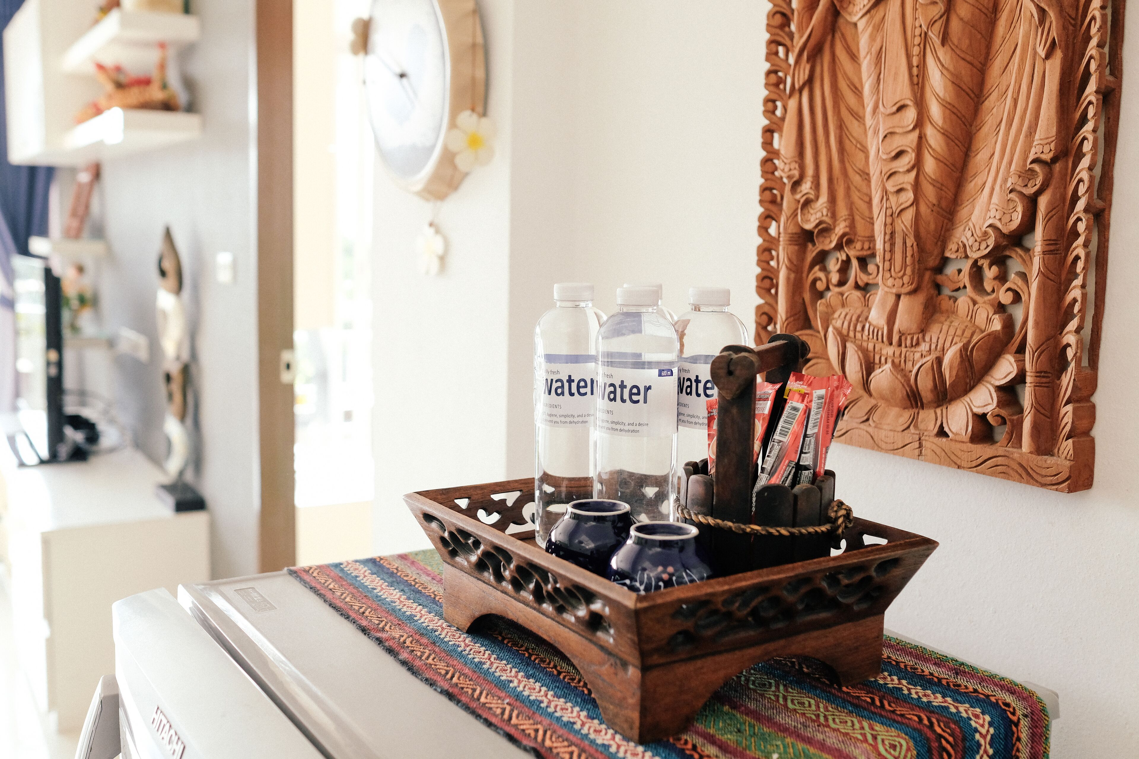 A wooden tray with water bottles and pens on a table, with a decorative wall carving in the background.