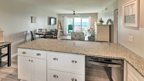 Una cocina con encimeras de granito, armarios blancos y lavavajillas. Hay una zona de comedor con mesa y sillas, y vistas al océano a través de amplias ventanas.
