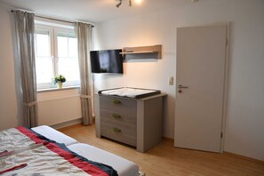 Una camera da letto con un letto, una cassettiera, una televisione a parete, una finestra con tende e una porta.