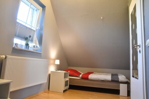 A small, compact room with a sloped ceiling, a single bed with red and white bedding, a small bedside table with a lamp, a window with blinds, and a radiator.