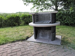 A circular outdoor grill with a stone base, surrounded by grass and a hedge.