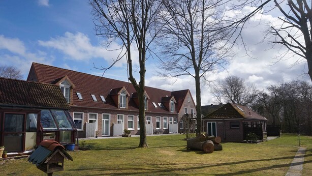 A residential building with a red-tiled roof, multiple windows, and a grassy yard with trees. - sparrow's nest - vikingerhof (Wittmund)