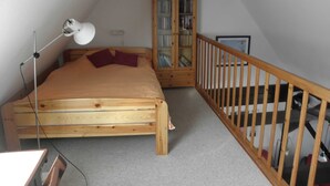 A bedroom with a wooden bed, a desk with a lamp, a bookshelf, and a staircase. - sparrow's nest - vikingerhof (Wittmund)