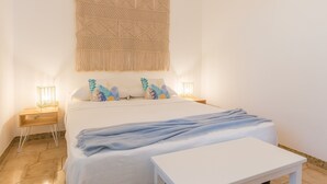 Une chambre à coucher avec un lit, des tables de chevet, une décoration murale en macramé et une petite table blanche.