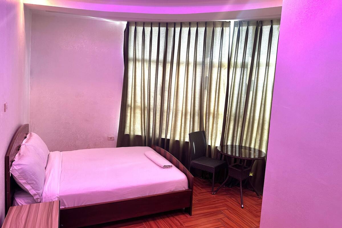 A hotel room with a bed, a small table with two chairs, and a window with curtains.