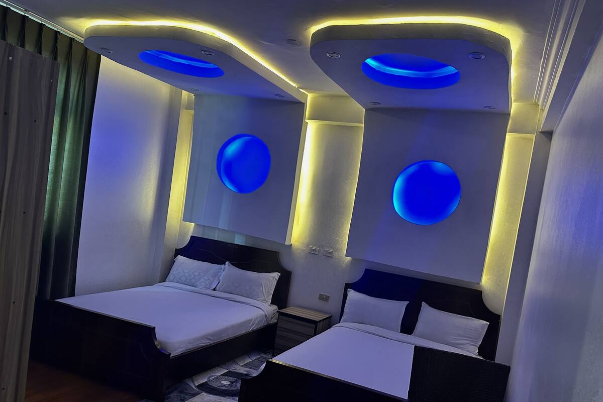 A modern hotel room with two beds, a large rug, and unique ceiling lighting.