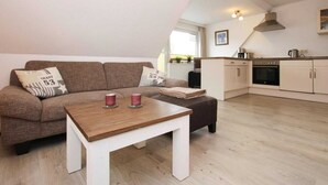 A cozy living room with a brown sofa, a wooden coffee table, and a kitchen area with white cabinets and a stove. - Ferienwohnung, St. Peter-Ording (St. Peter-Ording)