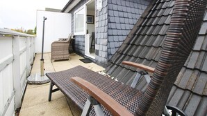 A covered outdoor seating area with a bench and a chair, a door leading to a room, and a white fence. - Ferienwohnung, St. Peter-Ording (St. Peter-Ording)