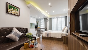 A modern hotel room with a leather sofa, a wooden coffee table with amenities, a bed with a TV, and a kitchen area in the background. - Narcissus Hotel and Apartment (Hanoi)