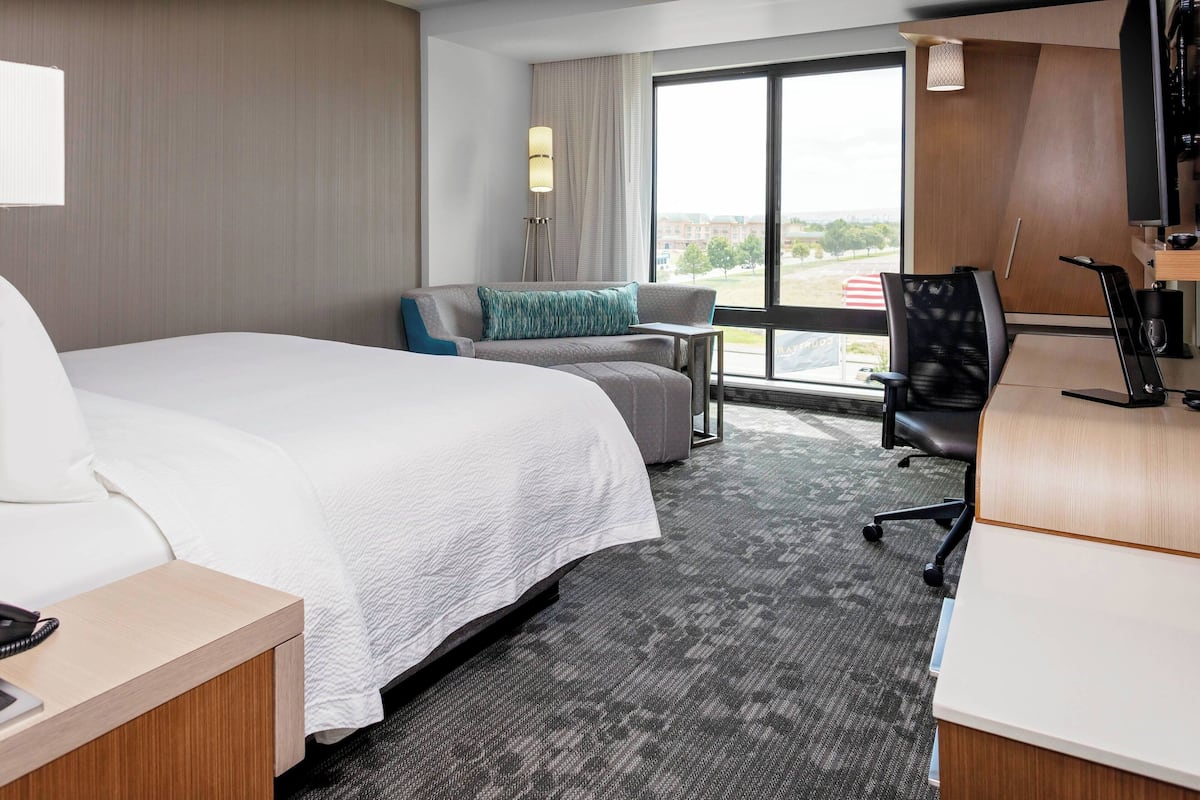 A hotel room with a large bed, a desk, a chair, and a view of the outdoors through a large window.