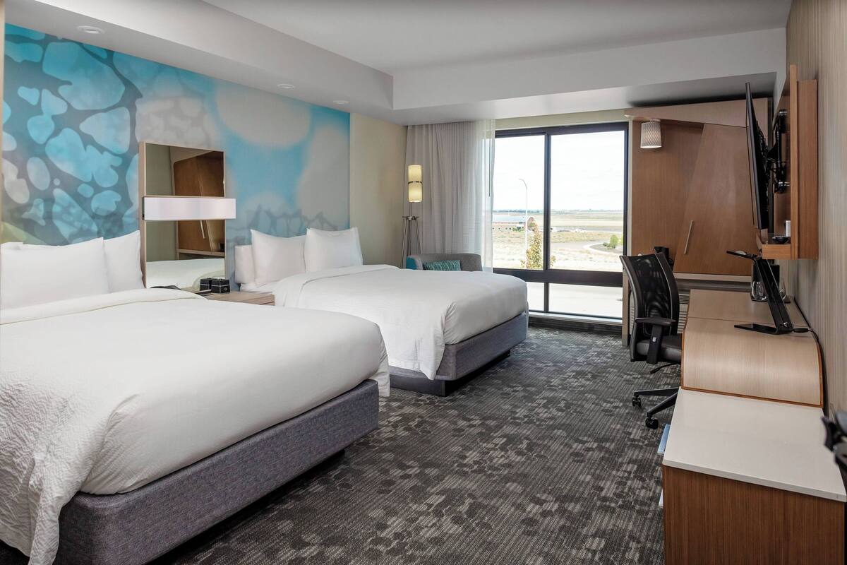 A hotel room with two beds, a desk, a chair, a lamp, and a large window with a view.