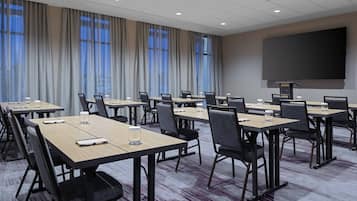 A conference room with multiple tables, chairs, a large screen, and windows with curtains.